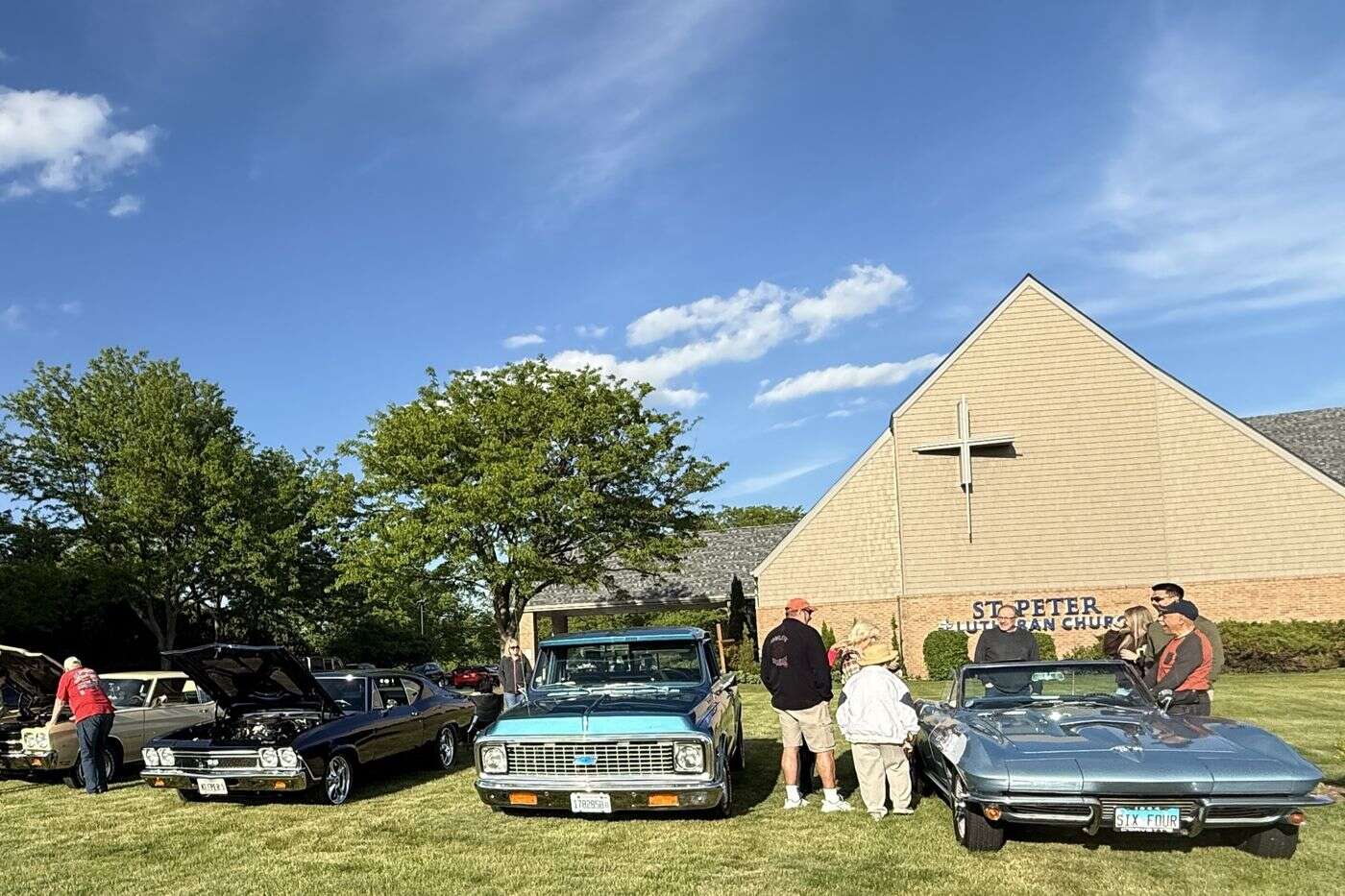 Classic cars and a vintage pickup truck parked on the grass in front of St. Peter Lutheran Church, with visitors walking between them on a sunny day.